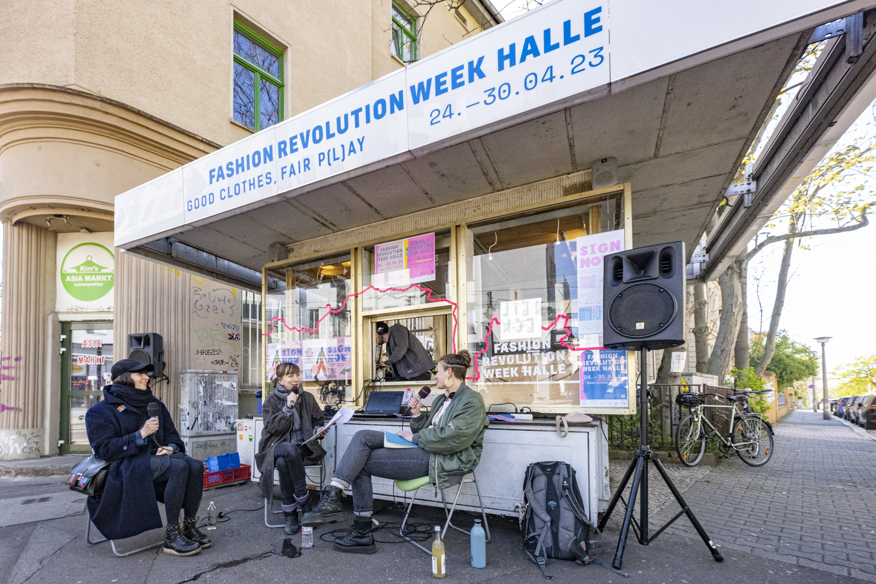 Ein öffentlicher Stand von Fashion Revolution. 3 Personen sitzen auf Stühlen davor, halten jeweils ein Mikrofon in der Hand und reden miteinander.